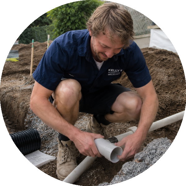 Plumber kneeling in a trench, connecting a gray PVC pipe at a construction site.