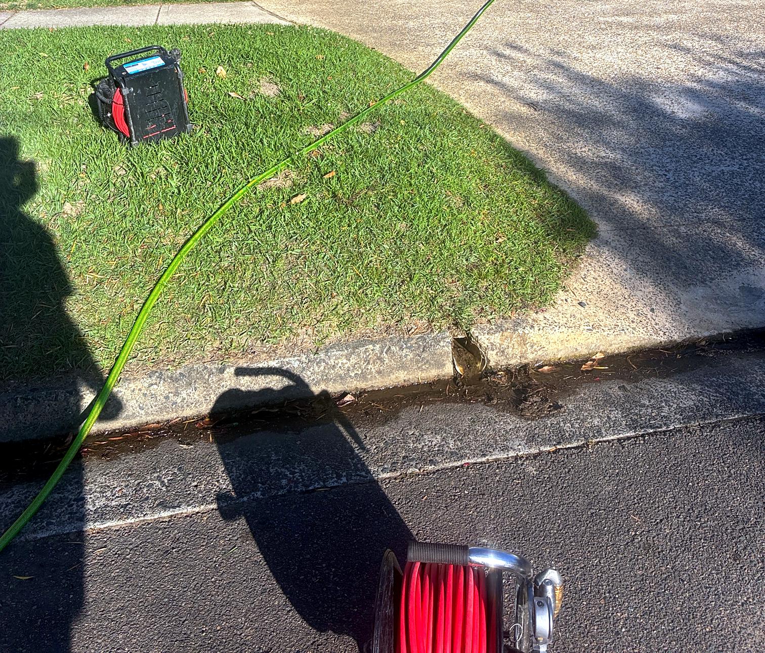 A drain camera unit on a grass verge with a hose running down to the stormwater outlet in the kerb