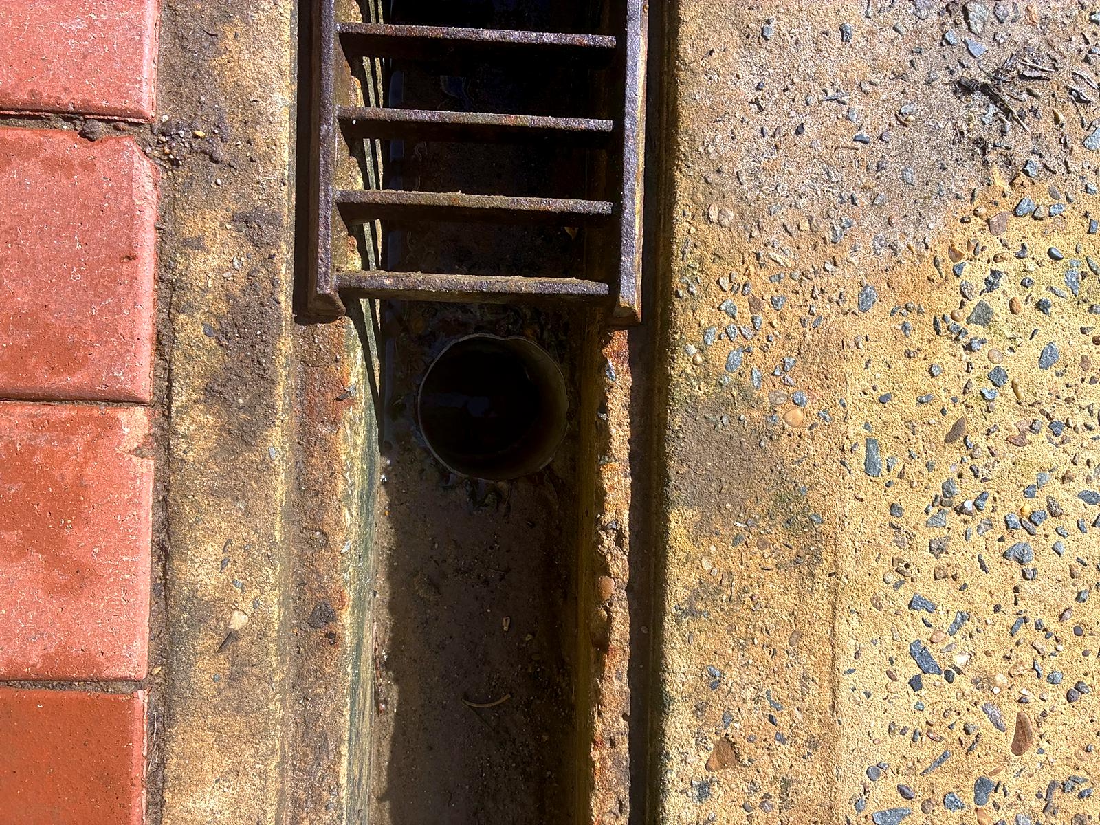 A strip drain with the grate lifted, showing the outlet pipe and silt in the base of the channel